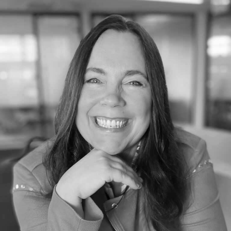 A woman with long dark hair smiles brightly at the camera, resting her chin on her hand. She is wearing a light-colored jacket and is indoors, with blurred windows in the background. The black-and-white photo gives an angus millar lecture 2025 vibe.