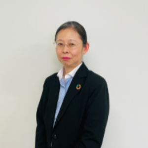 A woman with glasses and tied-back hair stands against a plain white background, wearing a black blazer over a light blue shirt and a white collar, her lapel adorned with a multicolored pin symbolic of sustainability events.