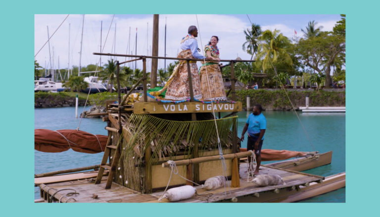 Two women in traditional dresses stand on a wooden raft named "Vola Sigavou," while a man in blue stands nearby. The raft is docked in a marina with sailboats, palm trees, and water in the background.