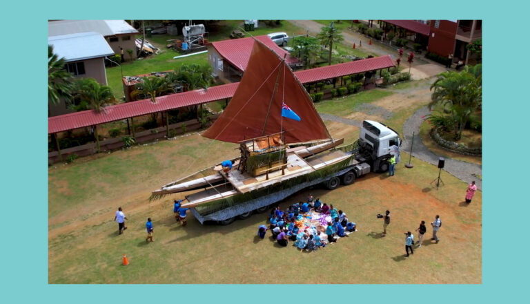 A large traditional outrigger canoe with brown sails is displayed on a flatbed truck in a grassy area. People are gathered sitting in circles nearby, with several buildings and palm trees in the background.