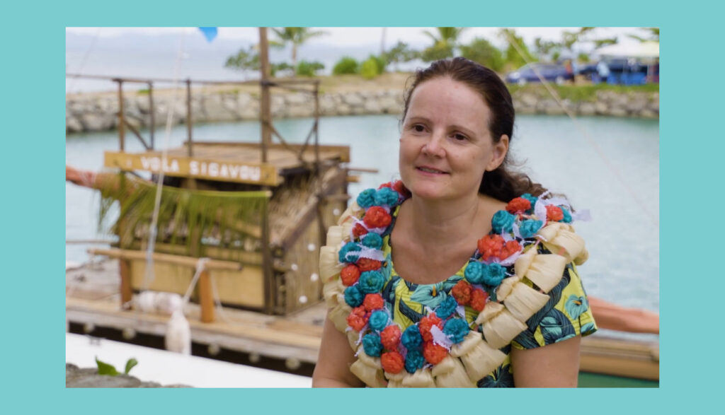 A woman wearing a colorful floral lei and a patterned dress sits near water with a traditional wooden boat and palm trees in the background, framed by a turquoise border.