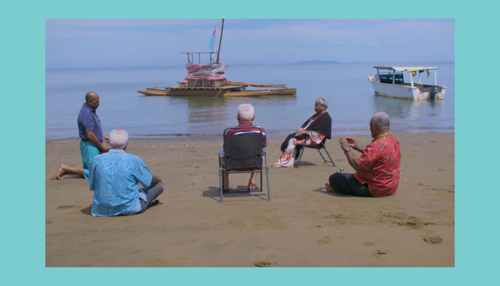 Five older adults sit and kneel on a sandy beach facing the sea, with two boats anchored near the shore under a clear sky. The scene is framed by a turquoise border.