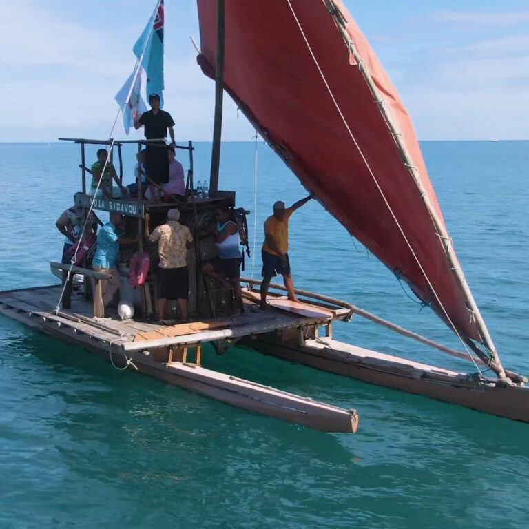 A group of people stands on a traditional wooden outrigger canoe with a large red sail, floating on calm blue ocean water under a clear sky.