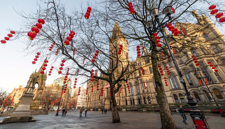 City square decorated with red and yellow Chinese lanterns hanging from trees, with a historic stone building and clock tower in the background. The scene suggests a festive celebration, possibly for Chinese New Year.