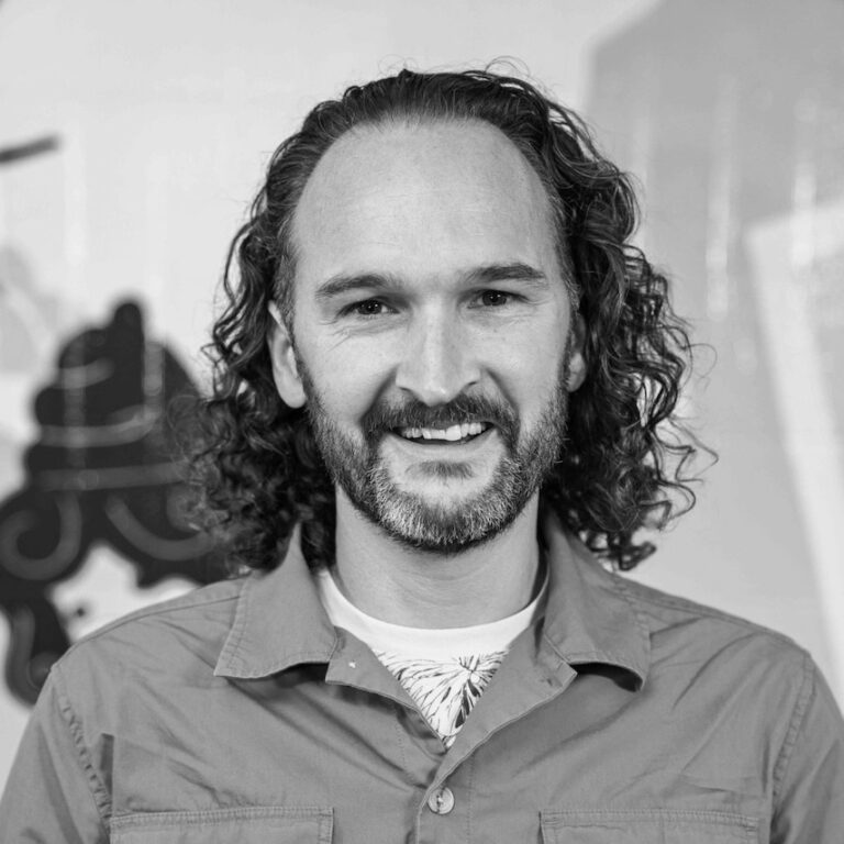Black and white photo of a smiling man with shoulder-length curly hair and a beard, wearing a button-up shirt over a T-shirt, standing in front of a patterned background—capturing the spirit of student innovation awards.