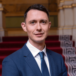 A man in a blue suit and tie stands indoors in front of a red-carpeted staircase with ornate railings, smiling slightly at the camera, possibly attending a sustainability event focused on the UN Sustainable Development Goals.