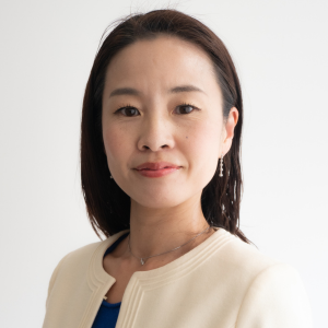 A woman with straight dark hair, wearing a cream blazer over a blue top, small earrings, and a delicate necklace, stands against a plain white background, looking confidently at the camera—ready to lead the next sustainability event.