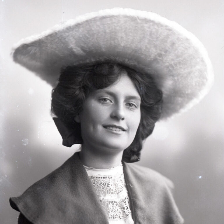 A woman with wavy dark hair smiles softly, wearing a large, wide-brimmed hat and a lace-collared blouse with a jacket. The image is black and white, with a soft, vintage look.