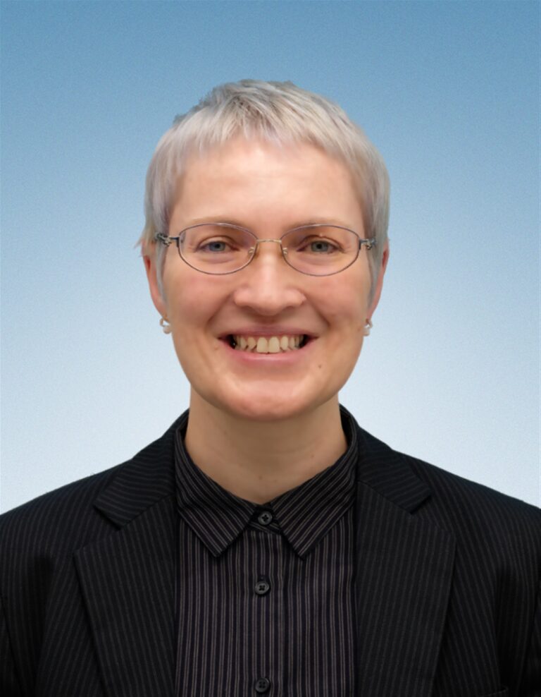 Smiling person with short gray hair and glasses, wearing a black pinstripe blazer over a dark shirt, stands in front of a light blue gradient background, ready to deliver the Edward Boyle Lecture.