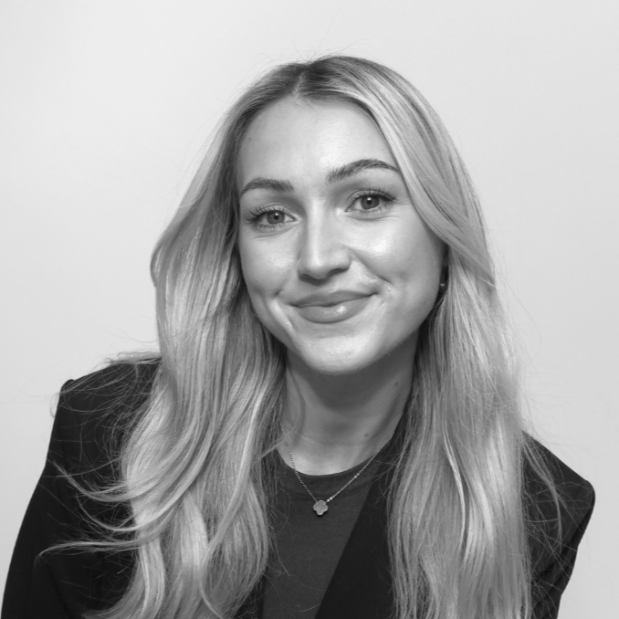 A young woman with long, wavy blonde hair smiles at the camera, radiating warmth and connection. She is wearing a dark blazer over a top and a small necklace. The black and white photo features a plain light background.