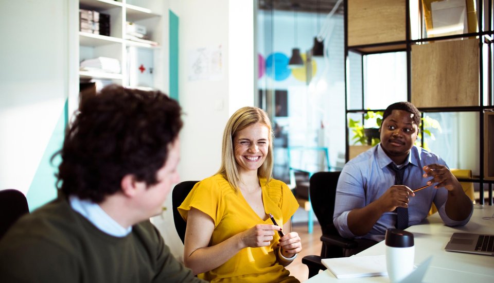 Three colleagues sit around a table in a bright office, smiling and talking after an engaging RSA webinar. A woman in a yellow shirt smiles at the man next to her, while another man sits across from them, holding glasses and smiling.