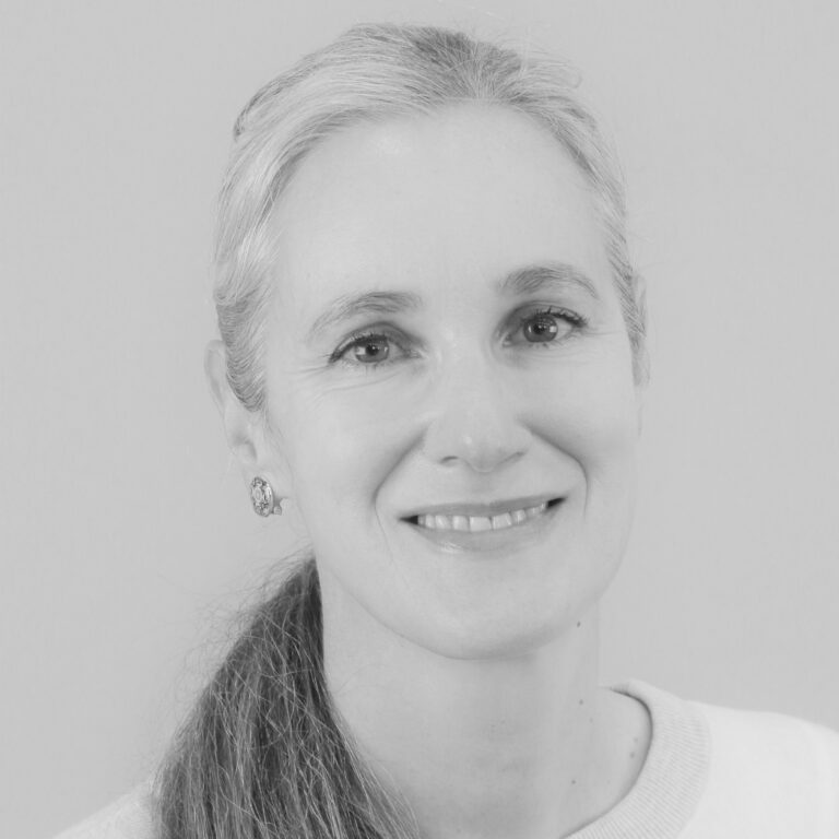 A woman with long, light-colored hair pulled back, wearing earrings and a light top, smiles at the camera against a plain background. The image is in black and white.