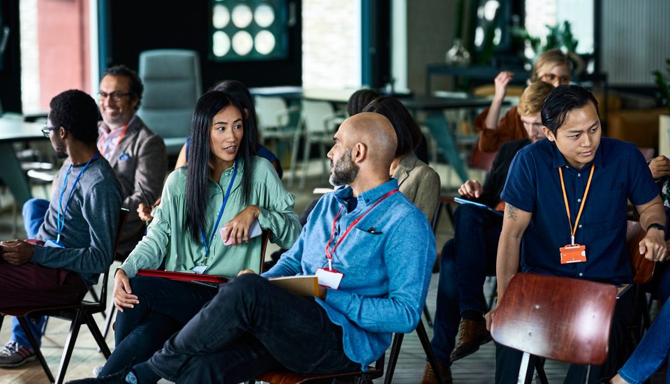 A group of people sit in chairs arranged in rows, wearing name badges and casually talking to each other in a modern indoor setting, suggesting a seminar or workshop environment.