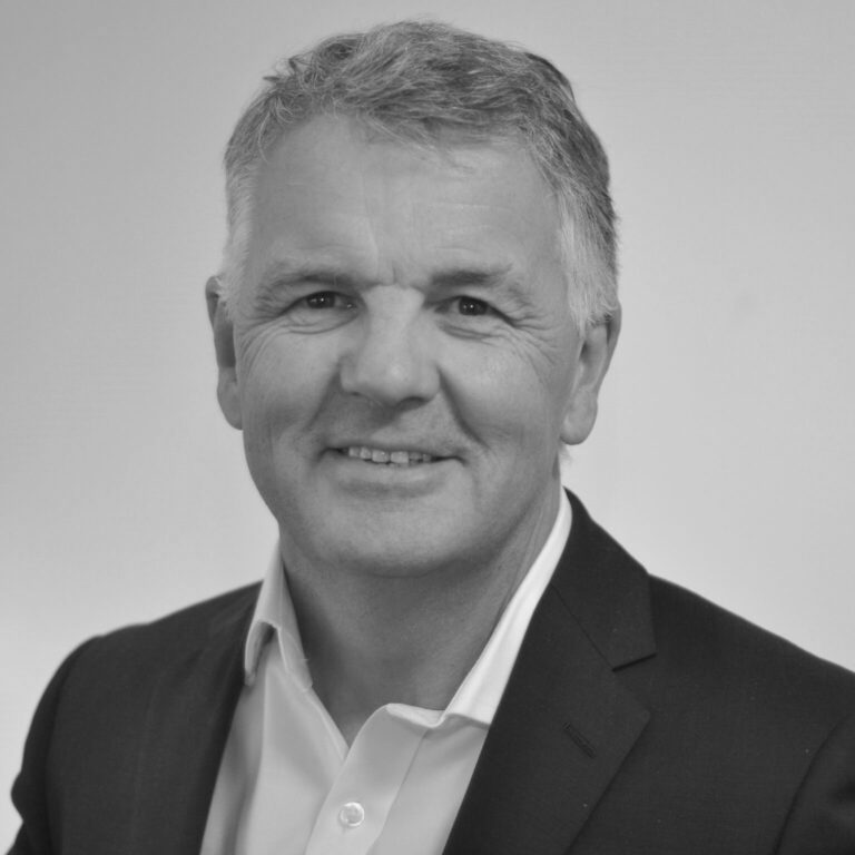 A middle-aged man with short grey hair, wearing a dark suit jacket and a white shirt, smiling at the camera against a plain light background. The image is in black and white.
