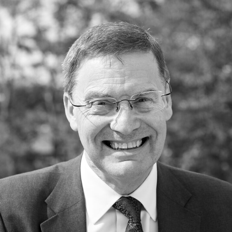 A middle-aged man with glasses and short hair, wearing a suit and tie, smiles broadly outdoors. The background is blurred with trees, and the image is in black and white.