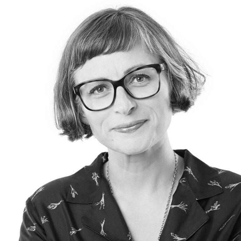 Black and white portrait of a smiling woman with short, straight hair and bangs, wearing large glasses and a patterned shirt, radiating warmth and connection against a plain white background.