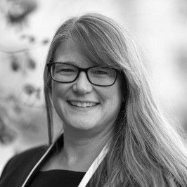 A woman with long, straight hair and glasses smiles at the camera. She is wearing a dark top and a light-colored lanyard. The background is blurred. The image is in black and white.