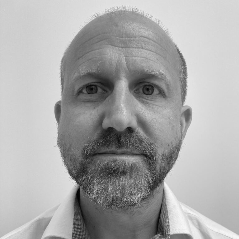 Black and white portrait of a middle-aged man with a beard and short hair, wearing a light-colored collared shirt, looking directly at the camera with a neutral expression, as part of the Edward Boyle Lecture series, against a plain background.