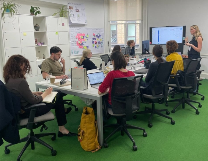 A group of people sit around two white tables in an office, listening to a woman presenting at a screen. Laptops, notebooks, and papers are on the tables; sticky notes cover a whiteboard in the background.