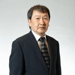A middle-aged man with short dark hair wearing a dark suit, white shirt, and striped tie stands against a plain, light gray background, ready to deliver the Edward Boyle Lecture.