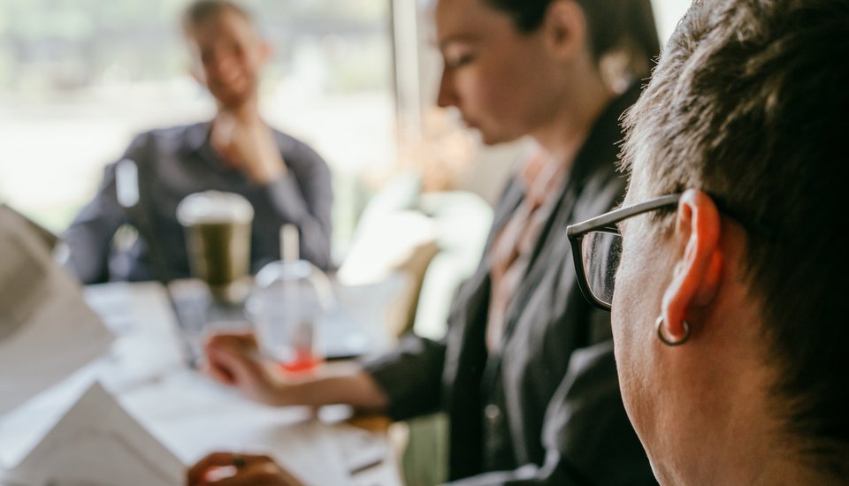 Three people dressed in business attire sit at a table, working with papers and drinks during york events. The foreground highlights one person wearing glasses and an earring, while two others are blurred in the background.
