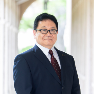 A middle-aged man wearing glasses, a dark suit, white shirt, and a red patterned tie stands in a sunlit corridor with arches and blurred greenery in the background, smiling gently at the camera—ready to host upcoming events on sustainability.