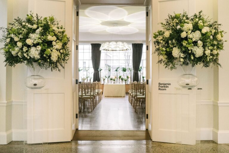 Double doors open to an elegant, well-lit banquet room with rows of wooden chairs and white floral arrangements; large flower displays flank the entrance on clear stands, and a sign reads "Benjamin Franklin Room.