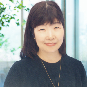 A woman with straight dark hair wearing a black top and a gold necklace smiles gently. She stands indoors in front of large windows with greenery visible outside, ready to attend events on sustainability and support the UN Sustainable Development Goals.