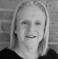 A woman with straight, light-colored hair and a friendly expression smiles at the camera. She is wearing a dark top, set against a brick wall in the background. The black-and-white image reflects the spirit of inclusive growth in the East Midlands.