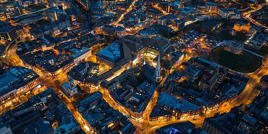 Aerial night view of a city with illuminated streets, buildings, and a central lit complex. Cars, parking lots, and a round park area with trees are visible among the warmly glowing urban landscape hosting lgbtqia events.