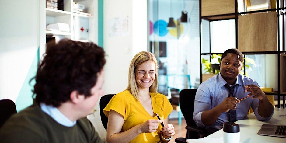 Three people sit at a table in a brightly lit office, smiling and talking about upcoming Birmingham events. One woman in a yellow shirt is in the center, holding a pen, while two men sit on either side, engaged in friendly conversation.