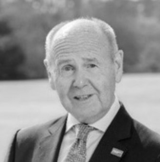 An older man with short, thinning hair, dressed in a suit and tie, is smiling slightly while standing outdoors in the East Midlands. The blurred black-and-white background suggests a grassy area with trees.