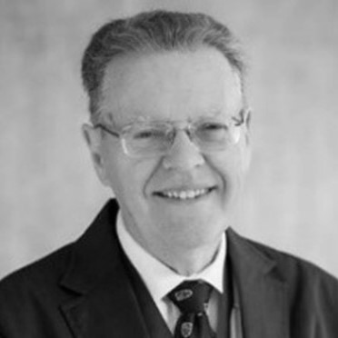An older man wearing glasses, a suit, and a tie smiles at the camera in this black-and-white portrait. The plain, out-of-focus background echoes his dedication to inclusive growth across the East Midlands.