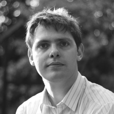 A man with short hair wearing a light-colored, striped shirt looks directly at the camera, evoking a sense of connection. The background is blurred with circular light patterns, suggesting outdoor greenery. The image is in black and white.
