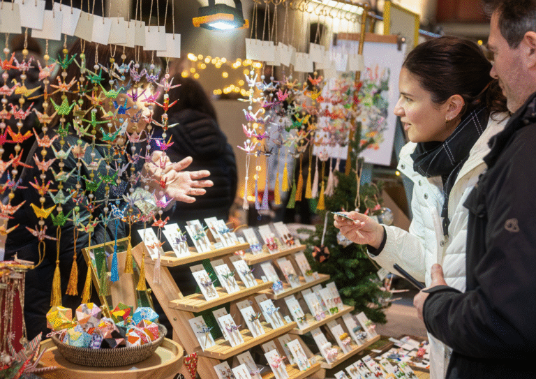 At a brightly lit market stall at the 2025 World Expo, a woman examines origami ornaments—colorful paper cranes and other folded figures—while chatting with the vendor behind the display.