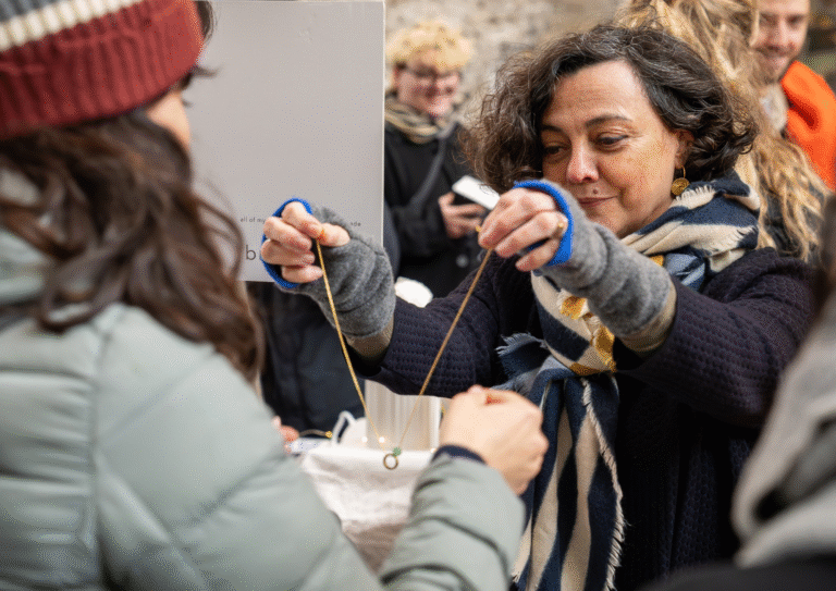 A woman in a scarf and gloves holds up a necklace, showing it to another person in a beanie and puffer jacket at an outdoor market, where smiles hint at excitement for the upcoming World Expo 2025 in Osaka.