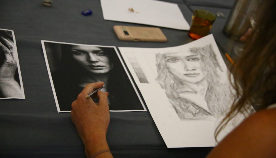 A person sketches a pencil portrait on white paper, referencing a black-and-white photo of a woman's face. Art supplies and a smartphone are visible on the table.