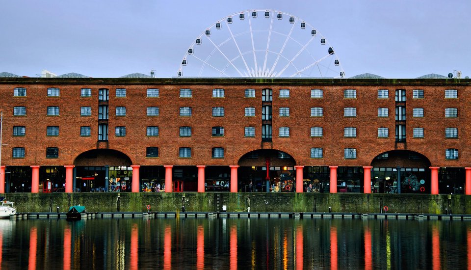 A large brick building with many windows stands by the water, its lower arches supported by red columns. A Ferris wheel rises in the background, and the building is reflected in the calm water below.