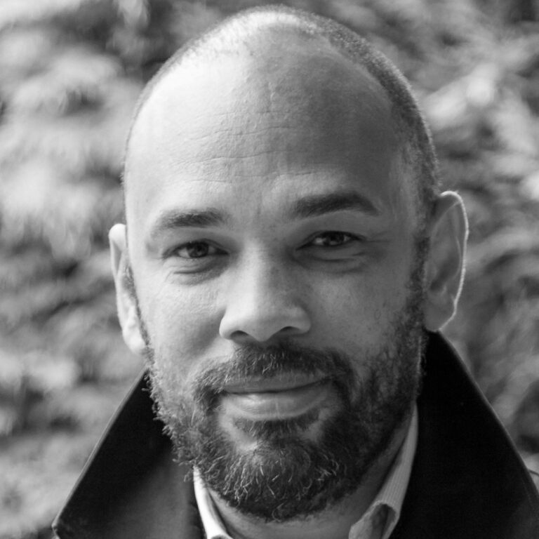 Black and white close-up portrait of a man with a beard and mustache, wearing a collared shirt and jacket, looking at the camera with a slight smile. A perfect profile image for freelancers in the creative industries, with blurred foliage in the background.