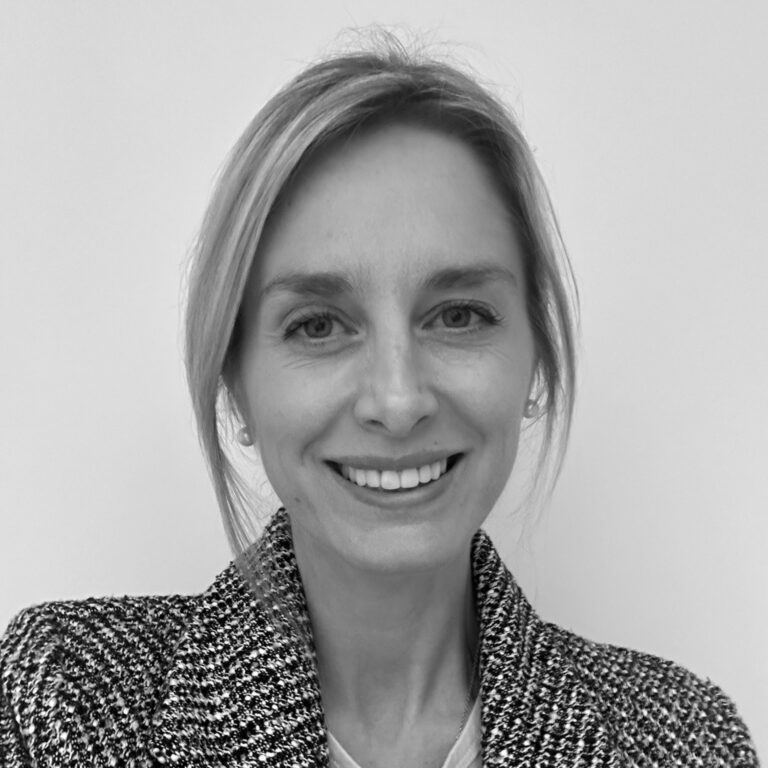 A woman with light hair tied back smiles at the camera. She is wearing a textured jacket and a light top, embodying the confident spirit of freelancers in the creative industries. The plain, light background complements this black-and-white portrait.