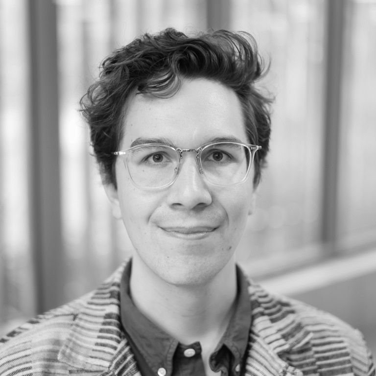 A person with short, curly dark hair and clear glasses smiles softly at the camera. Dressed in a patterned jacket over a collared shirt, they embody the style often seen in freelancers from creative industries. The blurred background hints at windows.