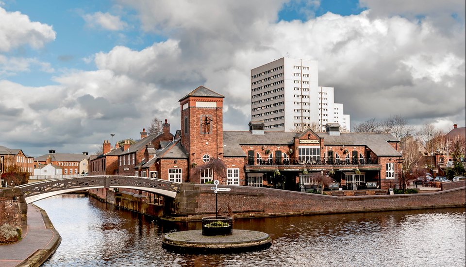 A canal with a small round island and a footbridge in front of historic red brick buildings. Modern white apartment towers rise in the background under a cloudy sky, blending old charm with the vibrant energy of events in Birmingham.