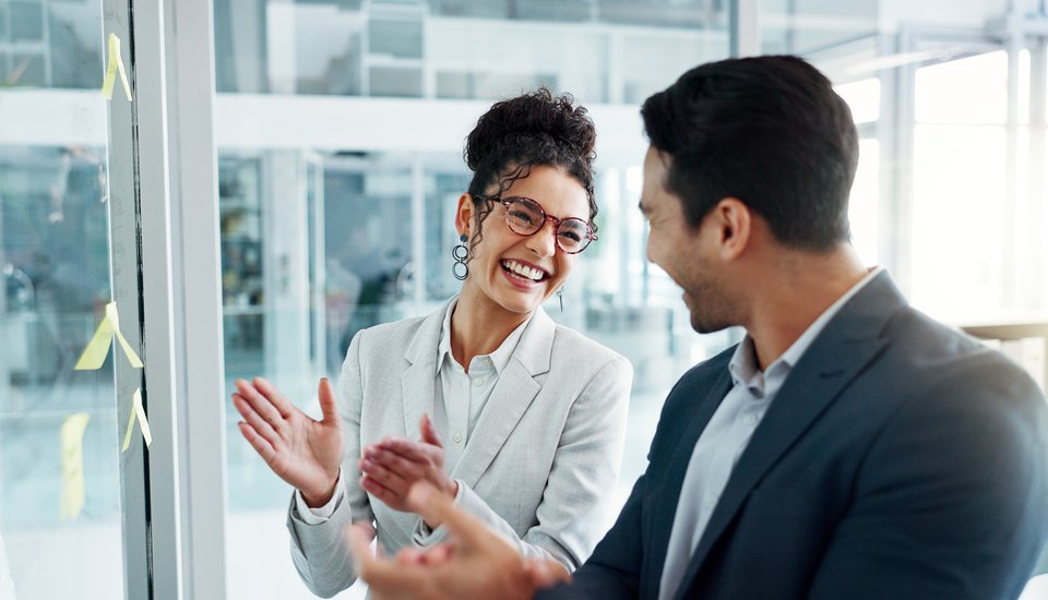 Two business professionals smiling and clapping in an office setting, celebrating or congratulating each other. Sticky notes are visible on the glass wall beside them.