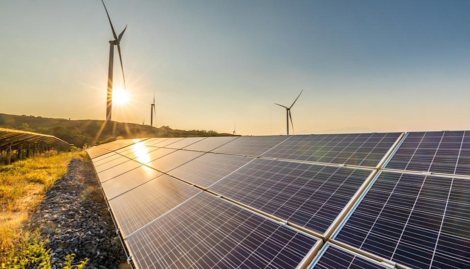 Rows of solar panels and tall wind turbines in a sunny, open landscape. The sun is low in the sky, casting warm light on the renewable energy installations and grassy terrain.