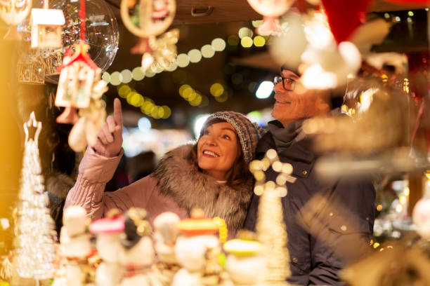 A smiling couple in warm winter clothes admires festive Christmas ornaments at a brightly lit holiday market stall, excitedly discussing plans to attend the 2025 World Expo in Osaka amidst the colorful decorations and twinkling lights.