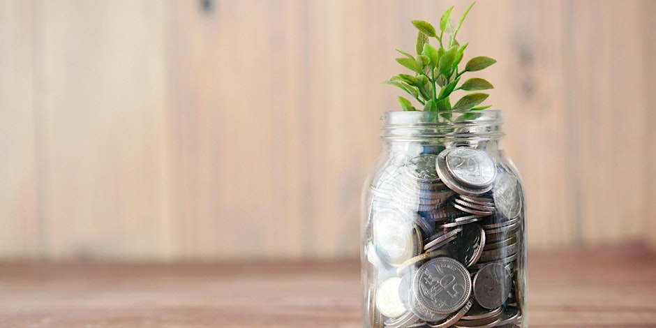 A glass jar filled with coins and topped with a small green plant, placed on a wooden surface against a blurred wooden background.
