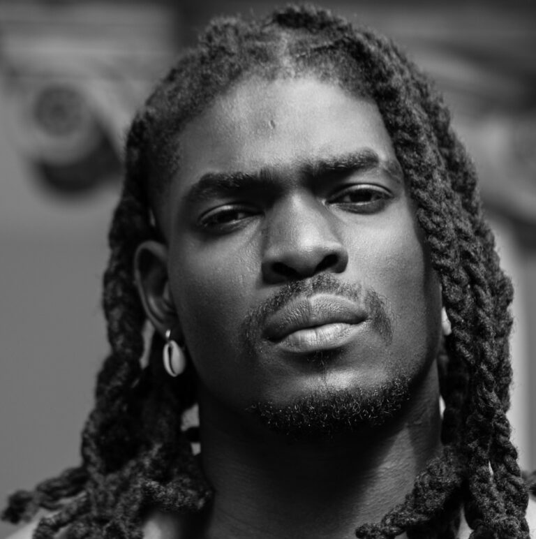 A serious-looking man with long, twisted dreadlocks and hoop earrings gazes directly at the camera in this black and white portrait, captured ahead of the New Black Film Collective film screening.