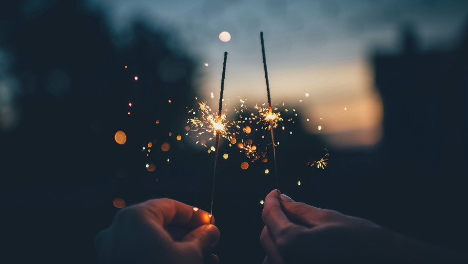 Two hands each hold a sparkling sparkler against a blurred, dark background with warm bokeh lights, celebrating community and togetherness in a festive atmosphere at dusk or night.