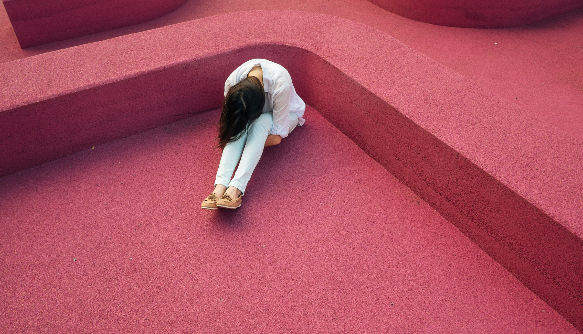 A person with long hair, wearing a white top and light pants, sits on a textured pink surface with their head down and arms wrapped around their knees, appearing to contemplate issues like community and gender equity.