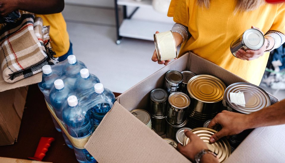 People pack canned food and bottled water into a cardboard box, with only their arms visible. A plaid blanket nearby suggests this could be a donation drive or disaster relief, perhaps part of charity-focused Birmingham events.